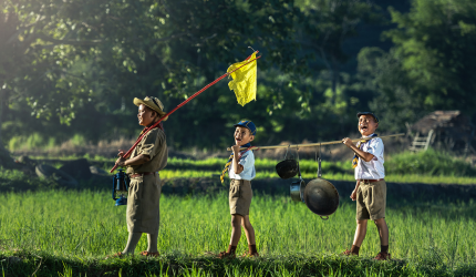 Three boy scouts walking through a grassy field carrying a flag and cooking pot