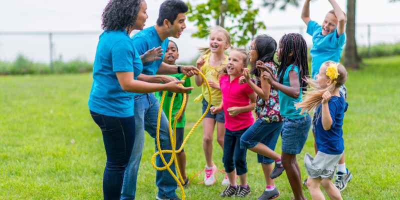 two adult camp counselors holding a jump rope with four kids watching